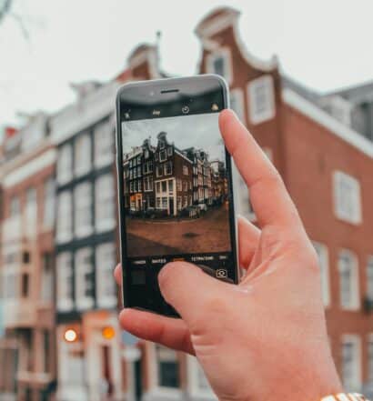 Uma mão branca segurando um celular na vertical para fotografar um dos prédios característicos de Amsterdam, para representar chip celular Amsterdam