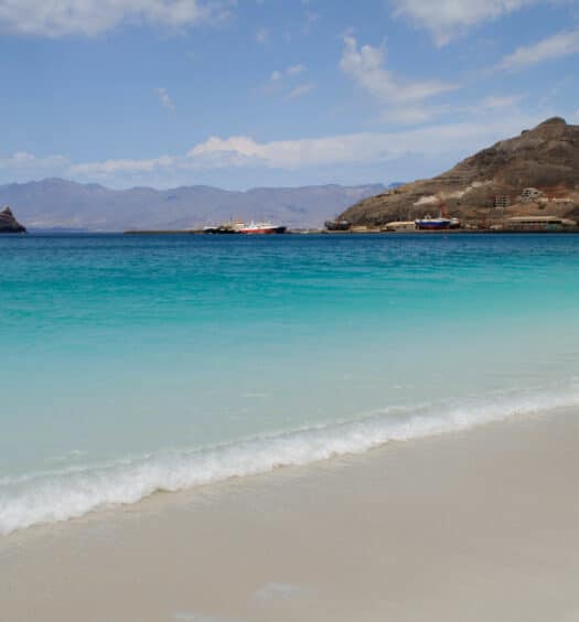 Praia com água num tom degrade azul, com montanha no fundo e barco navegando no mar. Imagem para ilustrar o post de chip celular Cabo Verde