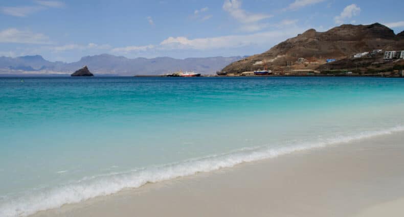 Praia com água num tom degrade azul, com montanha no fundo e barco navegando no mar. Imagem para ilustrar o post de chip celular Cabo Verde