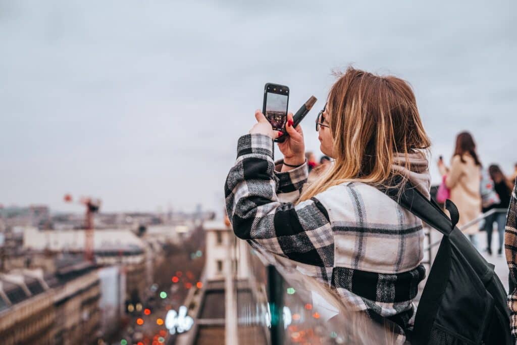 Uma moça jovem com uma casaco xadrez e uma mochila, ela está segurando um celular e está na cobertura de um prédio em Paris fotografando a cidade