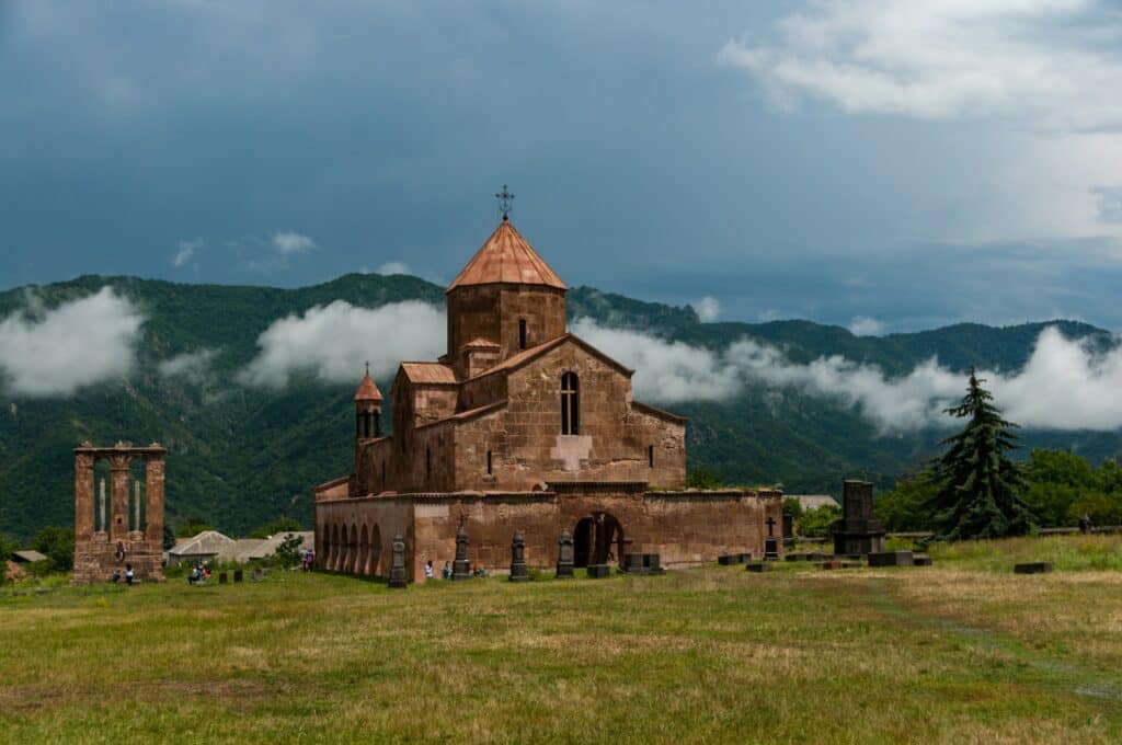 vista do Mosteiro de Odzun, Armenia, uma estrutura em pedra retos com uma abóbada mais alaranjada, com um gramado verde à frente, com algumas pessoas ao redor, ao fundo montanhas e o céu azul com neblina acima, para ilustrar o post de chip celular Ásia