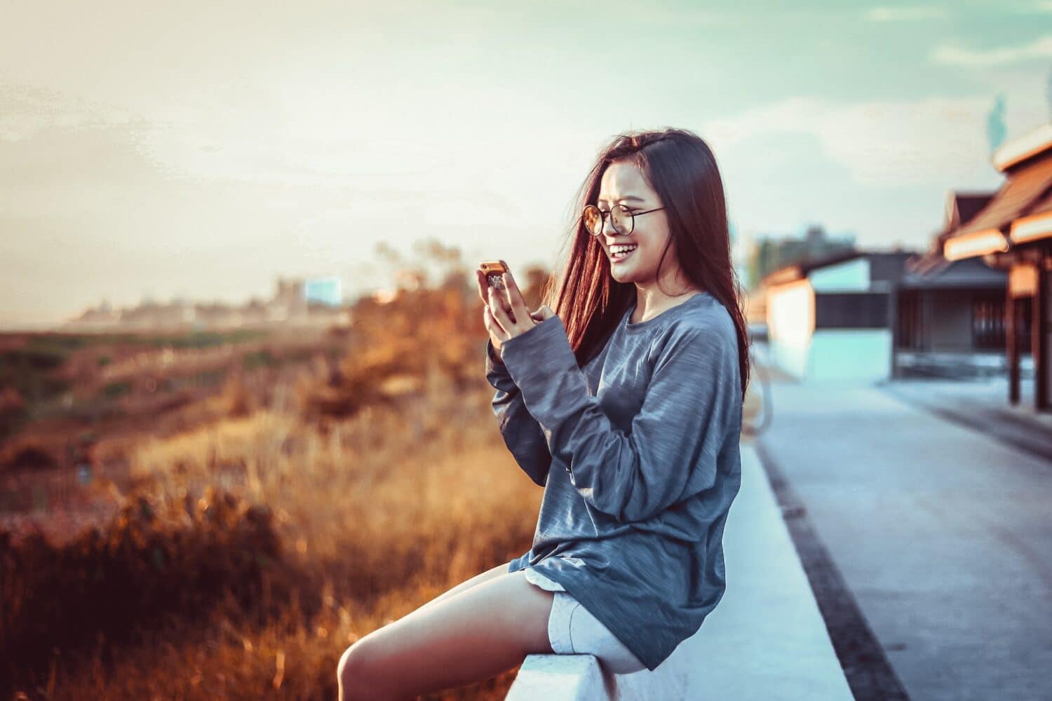 Mulher segurando um celular olhando para ele e sorrindo, com paisagem atrás. Ela está usando short jeans e blusa de manga longa cinza, imagem para ilustrar o post chip de celular Laos