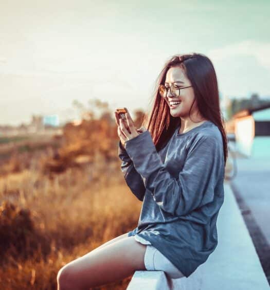 Mulher segurando um celular olhando para ele e sorrindo, com paisagem atrás. Ela está usando short jeans e blusa de manga longa cinza, imagem para ilustrar o post chip de celular Laos