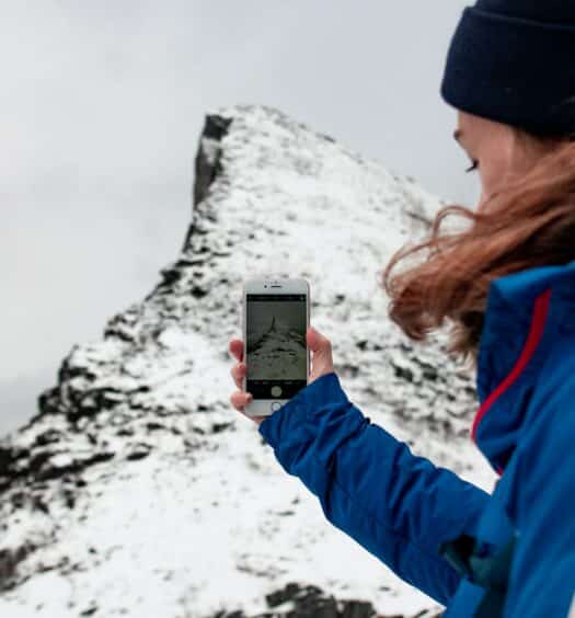 pessoa com roupas de frio e gorro segurando um celular e tirando foto de uma montanha cheia de neve para ilustrar o post de chip celular Noruega