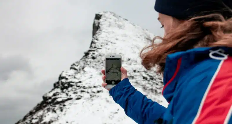 pessoa com roupas de frio e gorro segurando um celular e tirando foto de uma montanha cheia de neve para ilustrar o post de chip celular Noruega