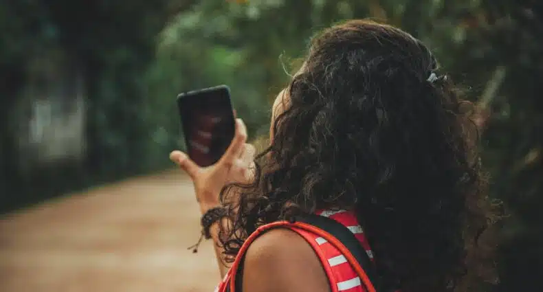 Uma mulher de cabelos enrolados e camiseta vermelha segurando o celular perto de um local com árvores por perto, para representar chip celular Sri Lanka