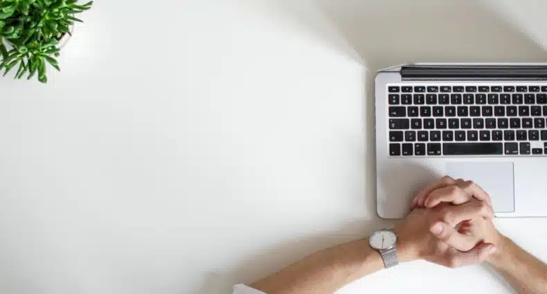 mesa branca com uma pequena planta verde aparecendo no canto esquerdo superior. No lado direito, visto de cima, é possível enxergar um notebook aberto com duas mãos masculinas brancas segurando uma na outra para representar o texto onde comprar um chip internacional