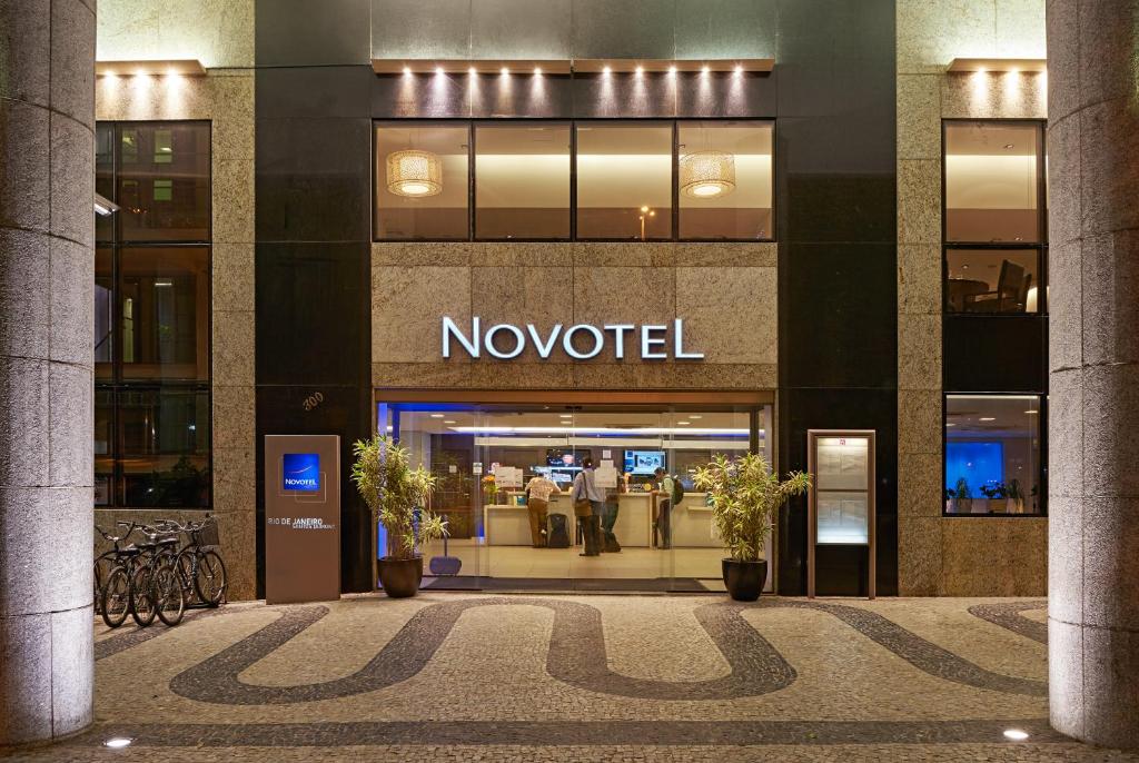 Entrada do hotel Novotel RJ Santos Dumont com duas pilastras de pedra, um bicicletário, janelas de vidro escuro e dois vasos de plantes