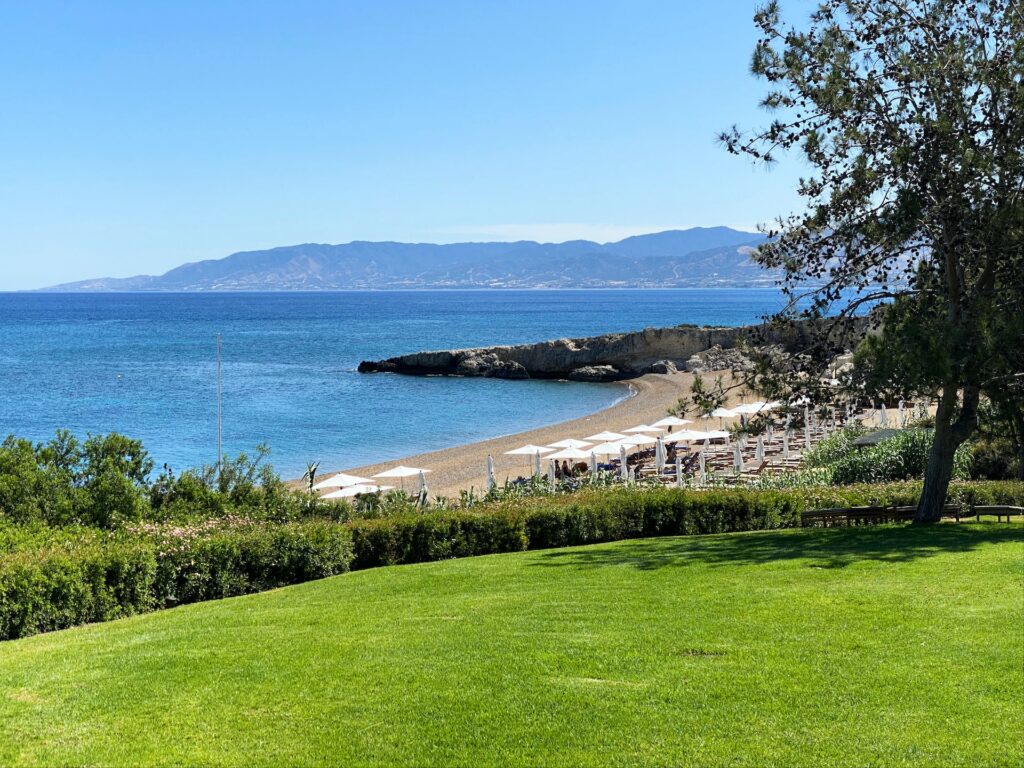 vista de uma praia em Pafos a partir de um campo de grama verde, a praia tem espreguiçadeiras e guarda-sol brancos com águas cristalinas azuladas e montanhas ao fundo com céu muito azul para ilustrar o post de chip celular Chipre