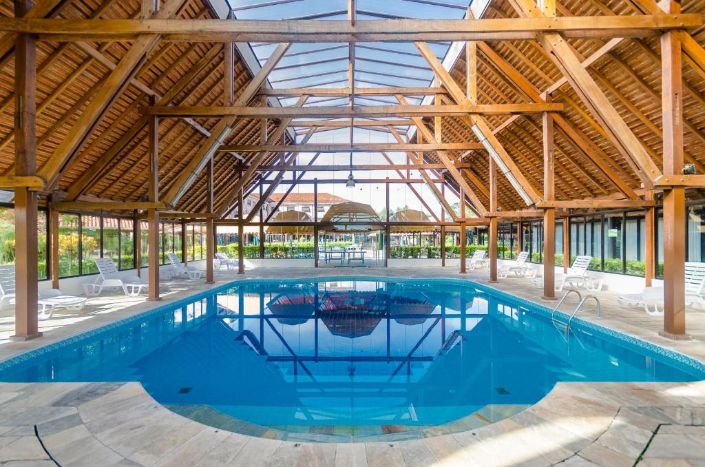 Área da piscina do Guararema Parque Hotel, com teto e estrutura de madeira, paredes de vidro, com vista dos jardins no lado de fora
