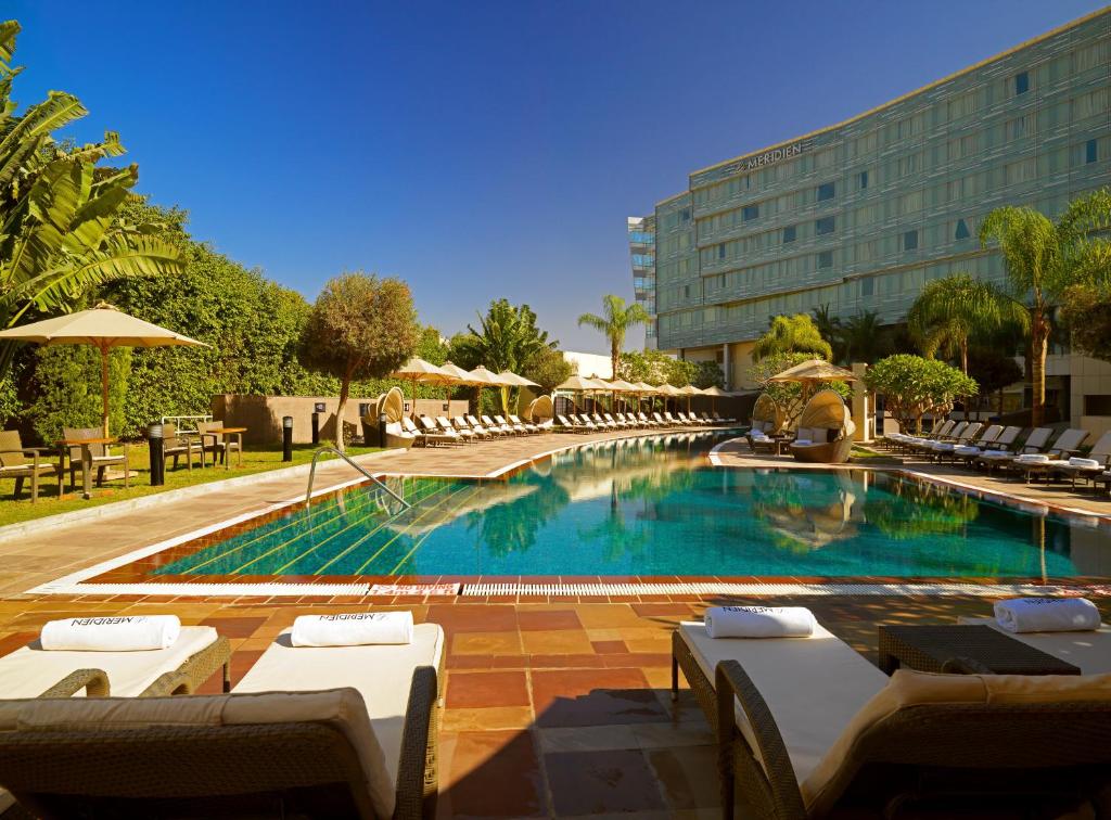 Vista da piscina do Le Meridien durante o dia com cadeiras em frente ao fundo da piscina com mais cadeiras na beira da piscina, coqueiros e jardim em torno da localidade. Representa Hotéis no Cairo.