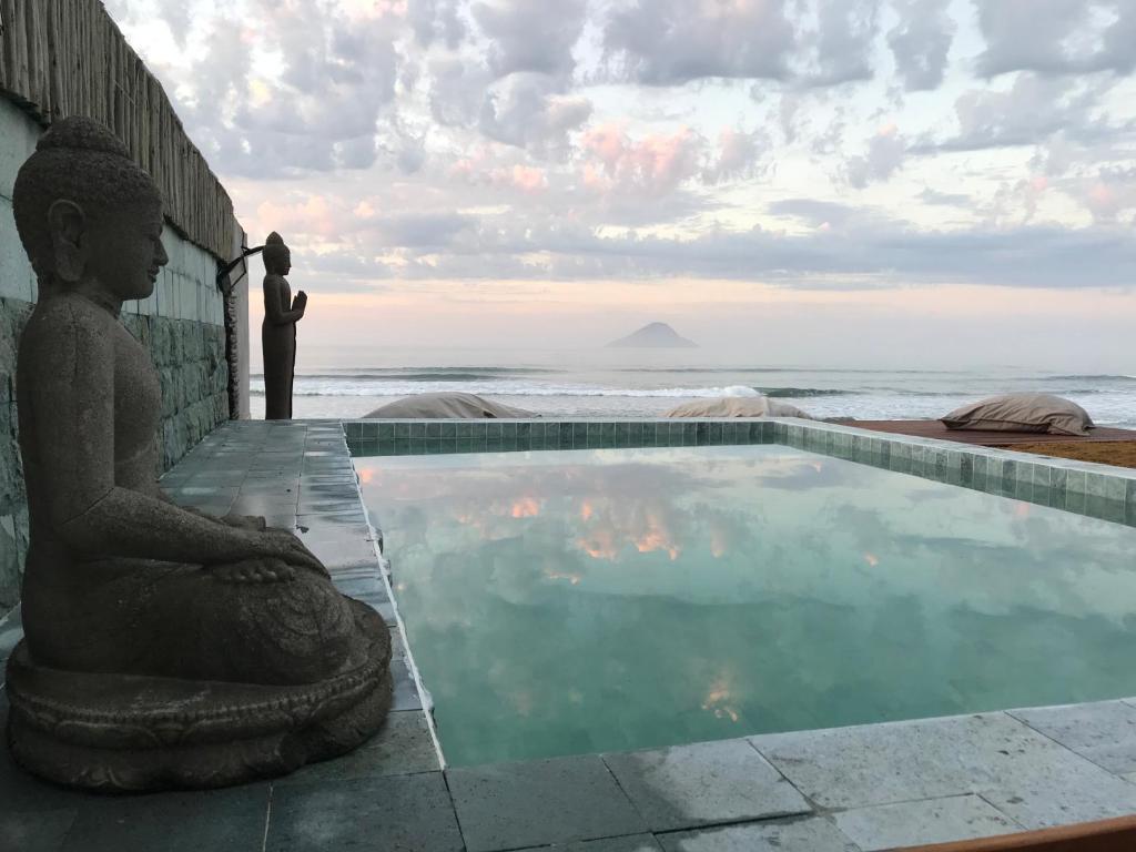 Vista da piscina da Casatua durante o dia com uma escultura indiana do lado direito e a frente da piscina vista para o mar. Representa pousadas em Camburizinho.
