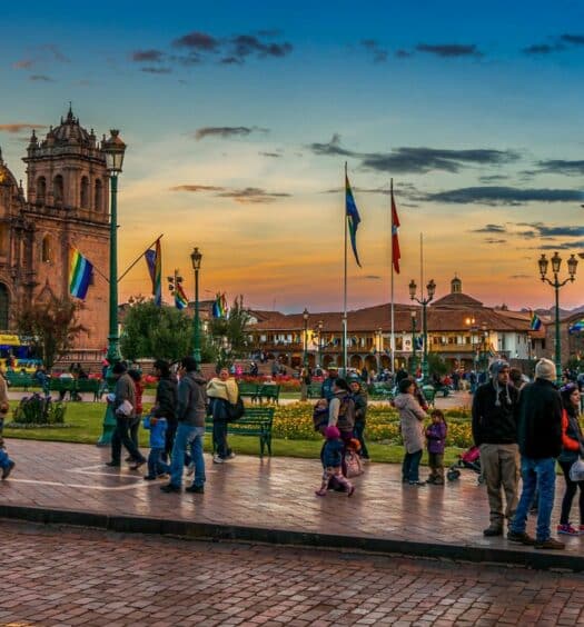 Várias pessoas passeando na Praça de Armas Cusco, sendo o local com dois prédios históricos, jardins e várias bandeiras com as cores do arco-íris. Imagem para ilustrar o post de chip de celular Cusco
