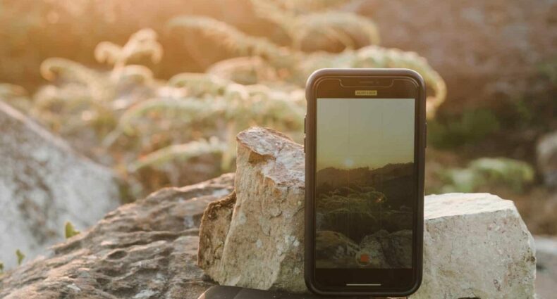 Celular com um aparelho ao lado, em cima de uma rocha e encostado em uma pedra, com paisagem e pôr do sol ao fundo. Imagem para ilustrar o post de chip de celular Nepal