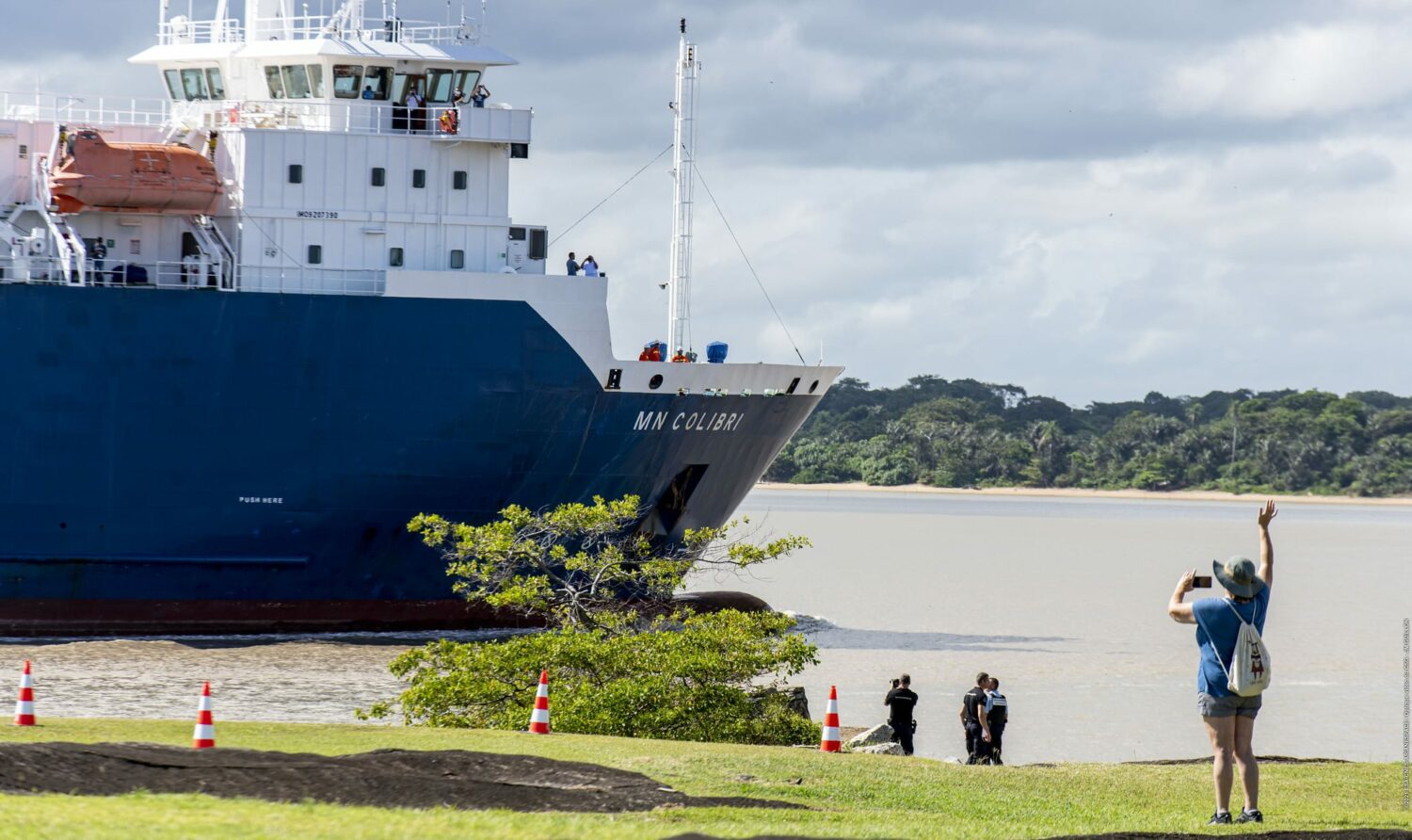 Porto com um navio azul e branco no mar, com pessoas na costa olhando e fotografando, na Guiana Francesa. Há também um gramado com uma árvore na costa do mar e montanhas ao fundo.