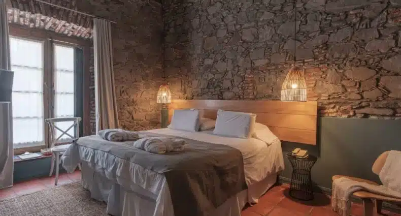 Quarto de pousada rústico com cama de casal, parede de pedra e piso cerâmico em tom terracota. É em Colonia del Sacramento.