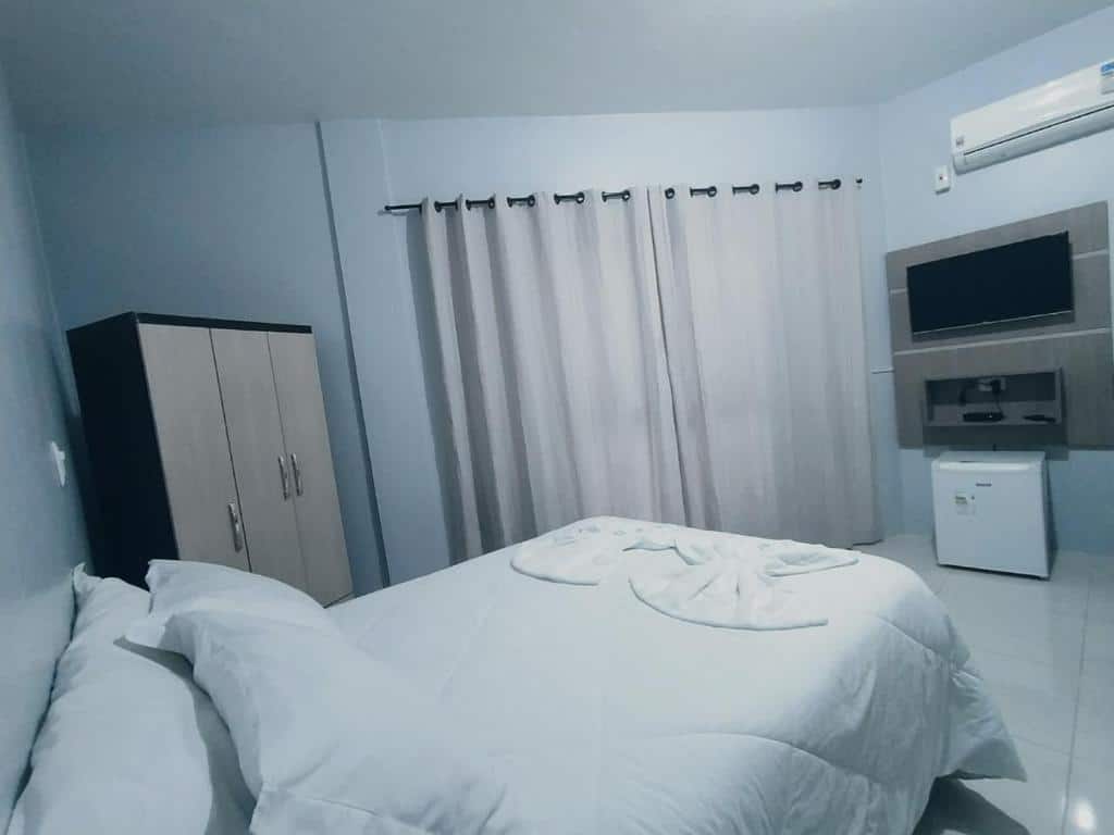 quarto da Pousada Aparados com uma cama de casal branca, um armário pequeno ao lado e uma televisão de tela plana em frente, logo abaixo de um ar-condicionado