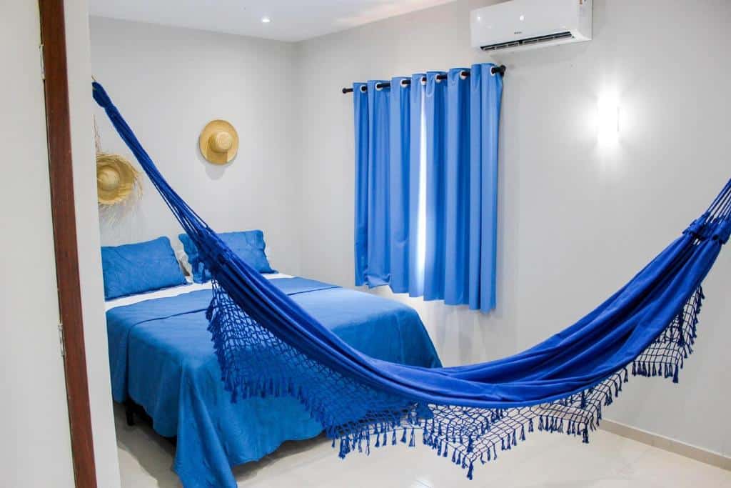 Quarto da Pousada Casa du Soleil, com cama com travesseiros e jogo de cama azul em cima, uma cortina azul na janela do lado direito e na frente tem uma rede do mesmo tom. Acima da cama tem dois chapéus de palha pendurados na parede