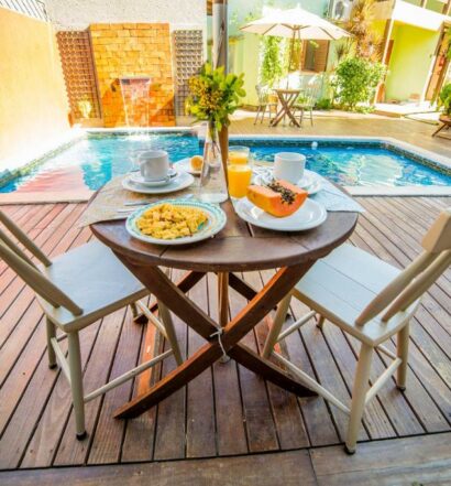Deck da Pousada do Capitão com uma piscina, uma mesa e duas cadeiras de madeira, sob à mesa há frutas e café, para representar pousadas na Praia do Campeche