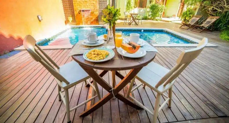 Deck da Pousada do Capitão com uma piscina, uma mesa e duas cadeiras de madeira, sob à mesa há frutas e café, para representar pousadas na Praia do Campeche
