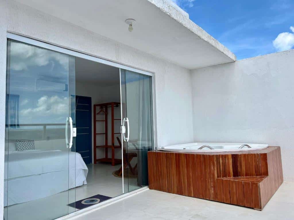 Quarto duplo deluxe da Pousada La Grécia, de 16 m², com uma banheira de hidromassagem redonda na varanda e uma porta de vidro dando acesso ao interior, que tem uma cama de casal, cadeira de madeira e cabideiro