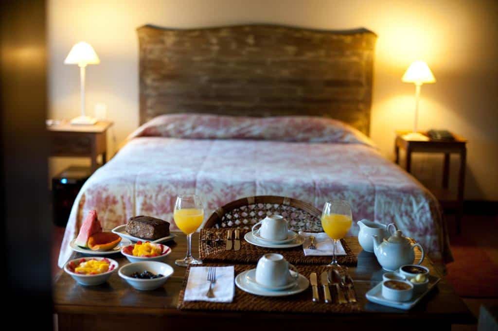 Quarto da Pousada Les Roches com uma cama de casal e uma mesa com um café da manhã disposto sob ela, com sucos, xícaras e bolos
