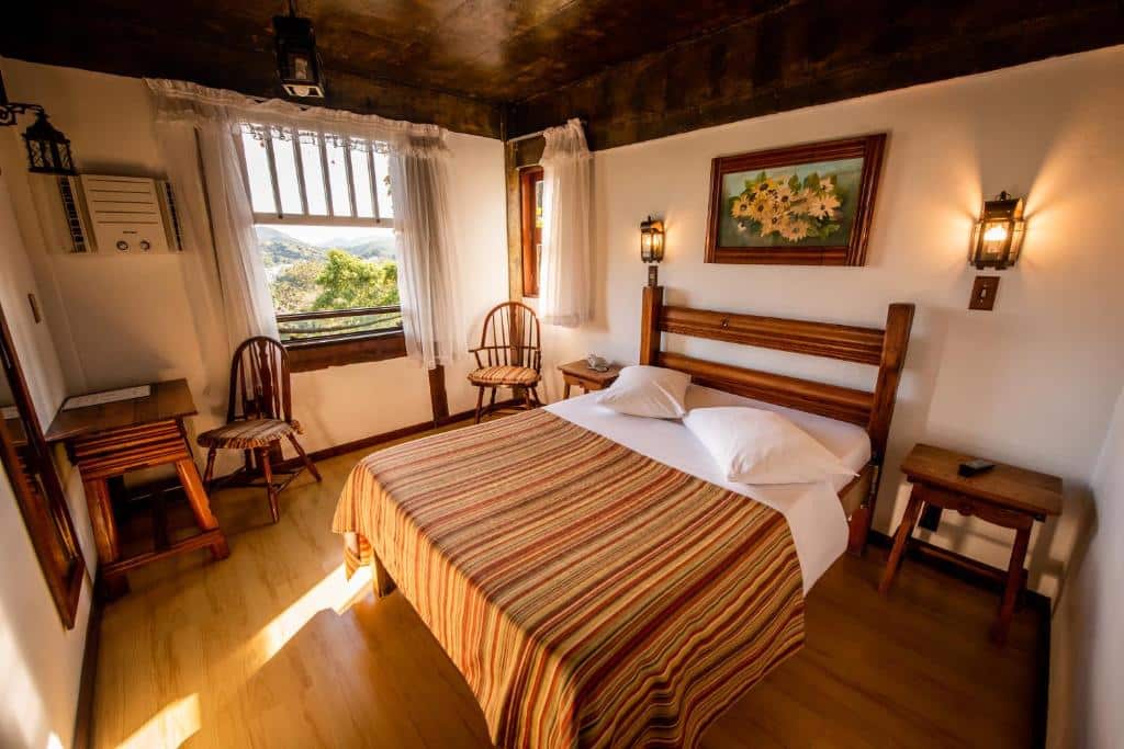 Quarto rústico da Pousada Monte Imperial com uma cama de casal, uma janela com vista para as montanhas, todos os móveis são de madeira, o espelho, as duas cadeiras, as mesinhas de cabeceira e uma mesa de trabalho