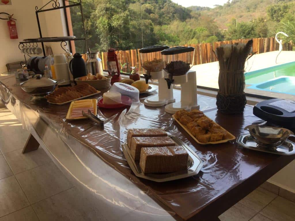 Mesa com café da manhã, parede de vidro e piscina ao fundo com vista da natureza
