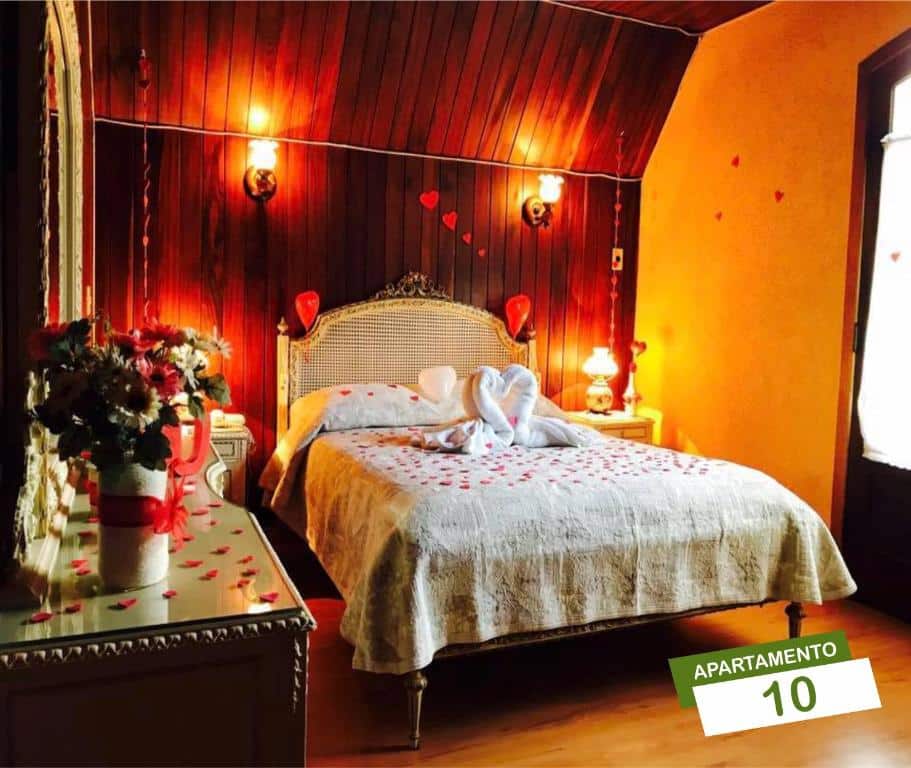 Quarto da Pousada Vale Verde com uma cama de casal, duas mesinhas de cabeceira com abajures, e uma cômoda com um espelho em cima, o ambiente tem pétalas de flores jogadas pelo chão