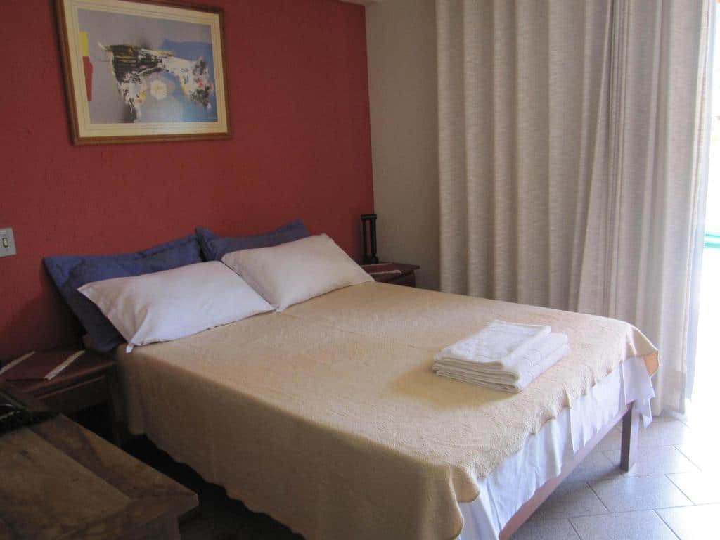 Quarto da Pousada Vento Sul com uma cama de casal, uma sacada com cortinas e um quadro na parede, para representar pousadas na Praia do Campeche
