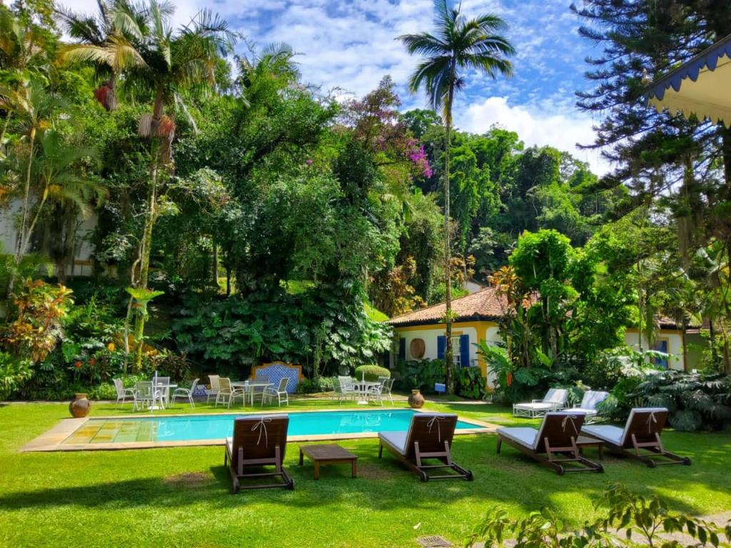 Jardim da Pousada Vila Brasil com muitas árvores ao redor, um amplo jardim com uma piscina, espreguiçadeiras de madeira e algumas mesas e cadeiras brancas