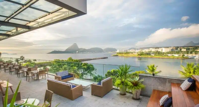Deck da cobertura do Prodigy Santos Dumont com sofás e poltronas, além de alguns vasos de plantas, com uma vista direta para o mar, cidade e muitas montanhas, para representar hotéis perto do Aeroporto Santos Dumont
