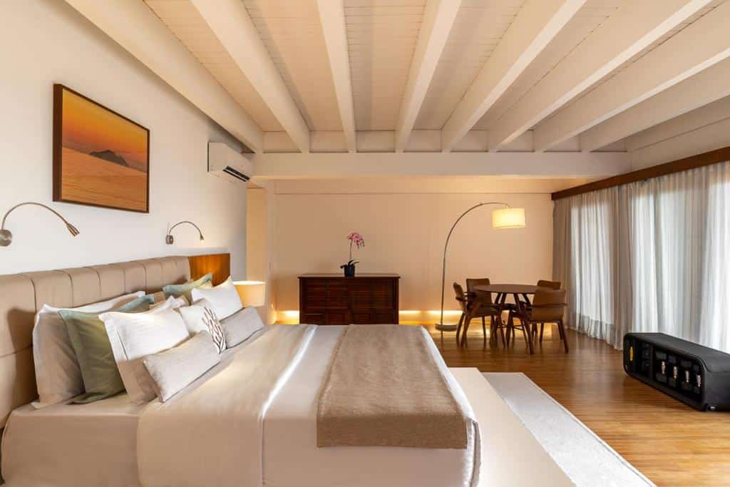 Quarto do A Concept Hotel & Spa com uma cama de casal, uma cômoda de madeira com um vaso de flor sob ela, uma mesa com quatro cadeiras, tudo em estilo minimalista e em tons de bege e madeira