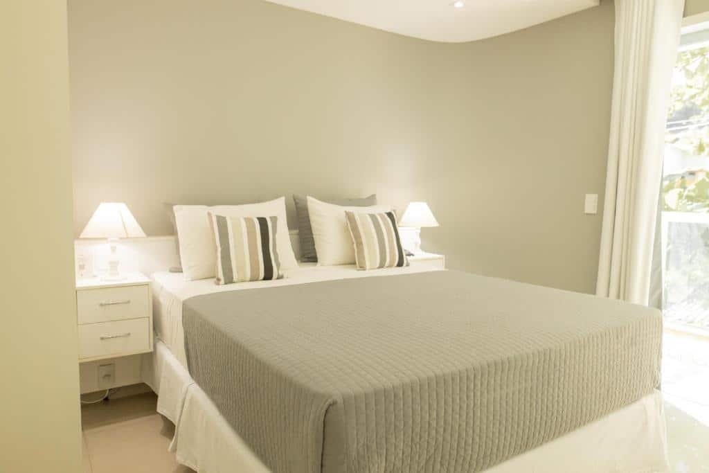 Quarto no Angra Beach Hotel com uma cama de casal com travesseiros e almofadas, duas mesinhas de cabeceira com abajures e uma sacada com cortinas do lado direito