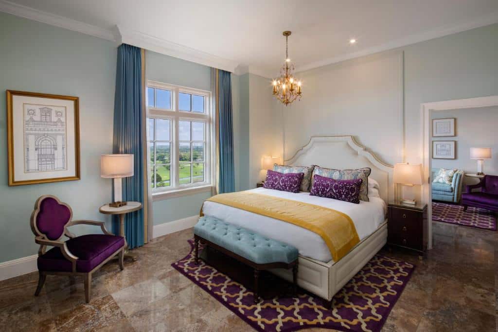 Quarto do Biltmore com cama de casal, duas cômodas do lado da cama com luminária, janela com cortinas azuis do lado esquerdo e a frente a cama poltrona roxa.