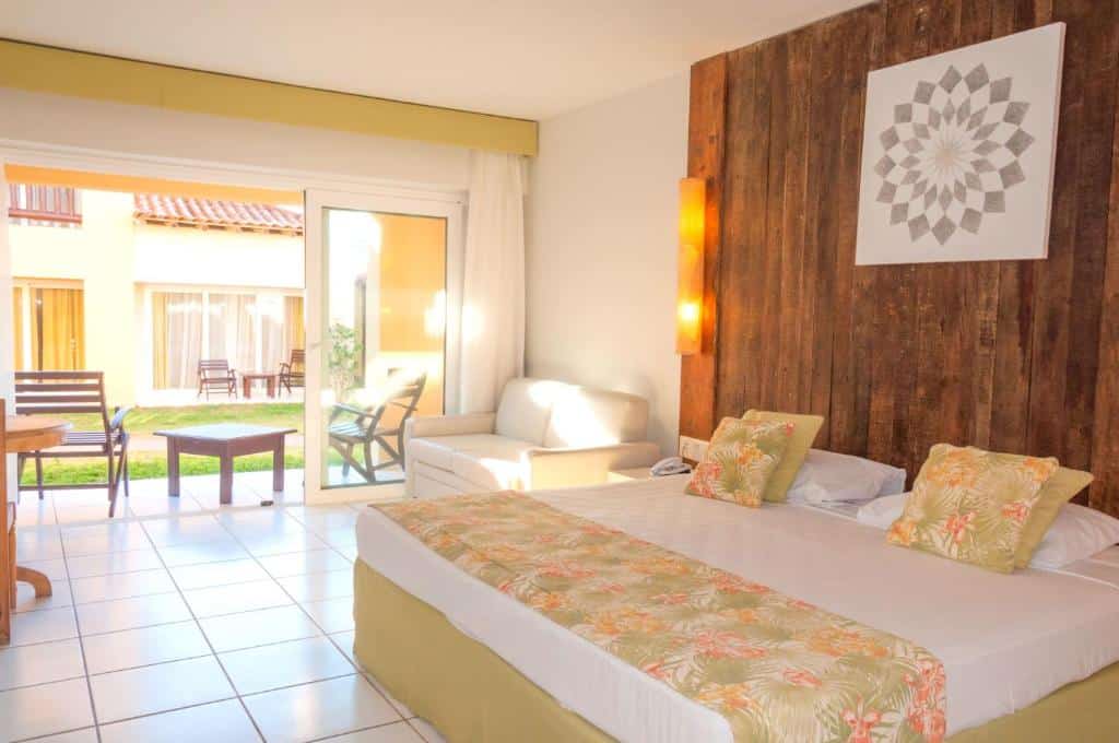 Quarto do Búzios Beach Resort com uma varanda térrea que leva para o jardim, do lado de dentro há um sofá de dois lugares, uma cama de casal e uma cabeceira de madeira rústica na parede atrás da cama com iluminação indireta