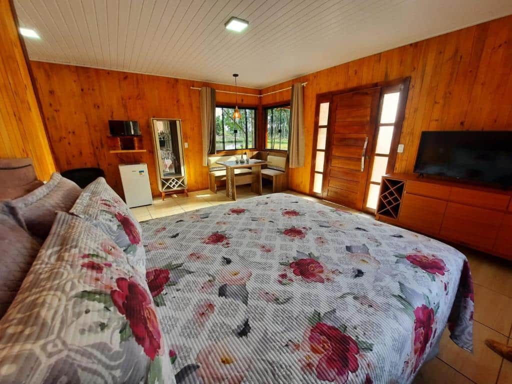 vista interna do quarto com uma cama de casal, mesa e cadeiras de canto com janelas amplas, frigobar e televisão de tela plana