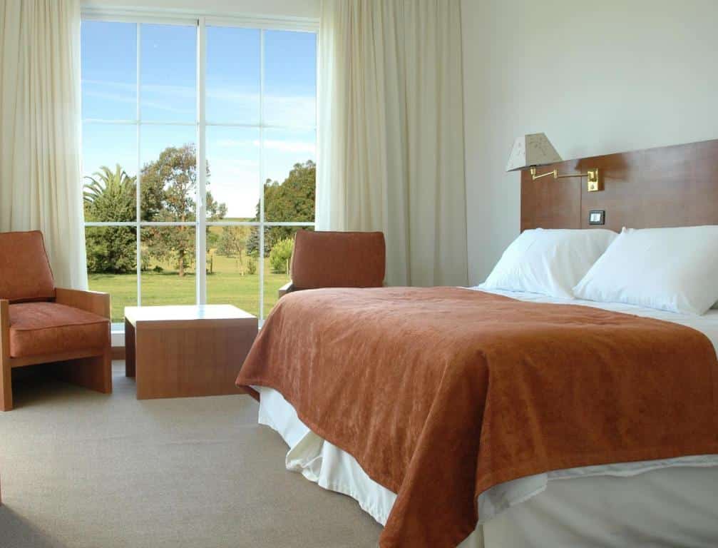 Quarto do Casa del Sol Hotel & Restaurante com uma cama de casal, uma mesinha de madeira, duas poltronas e um janela que vai até o chão com cortinas