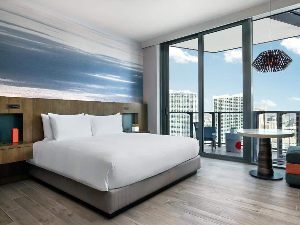 Quarto do East Miami com cama de casal ao centro do quarto, duas cômodas de madeira, mesa redonda do lado esquerdo com portas de vidro ao lado com varanda.