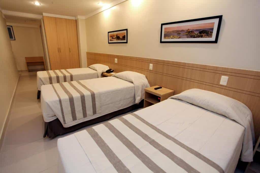 Quarto do Fluminense Hotel com três camas de solteiro, um armário de três portas, duas mesinhas de cabeceira e dois quadrinhos na parede, para representar hotéis perto do Aeroporto Santos Dumont