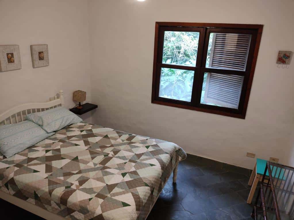 Vista do quarto da Casa aconchegante em Guaeca com cama de casal, luminária do lado esquerdo com janelas de madeiras.