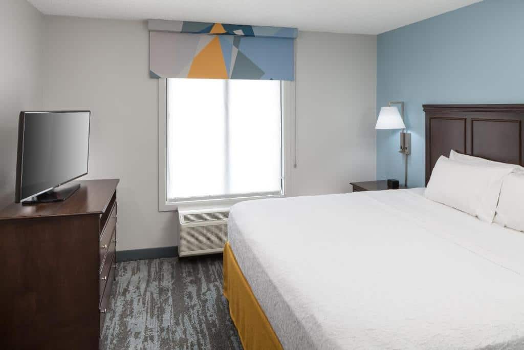 Quarto do Hampton Inn & Suites Miami-Doral Dolphin Mall  com cama de casal, cômoda do lado esquerdo, cômoda de madeira em frente a cama com TV.