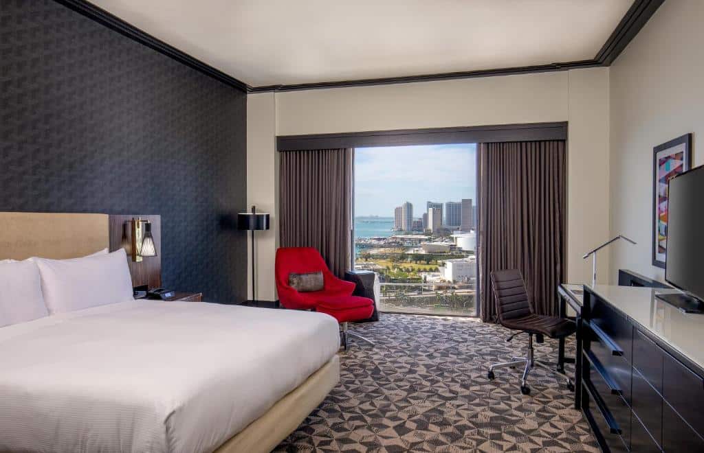 Quarto do Hilton Miami Downtown com cama de casal, cômoda com luminária ao lado esquerdo da cama, do lado esquerdo poltrona vermelha e em frente a cama cômoda com TV e mesa de trabalho.