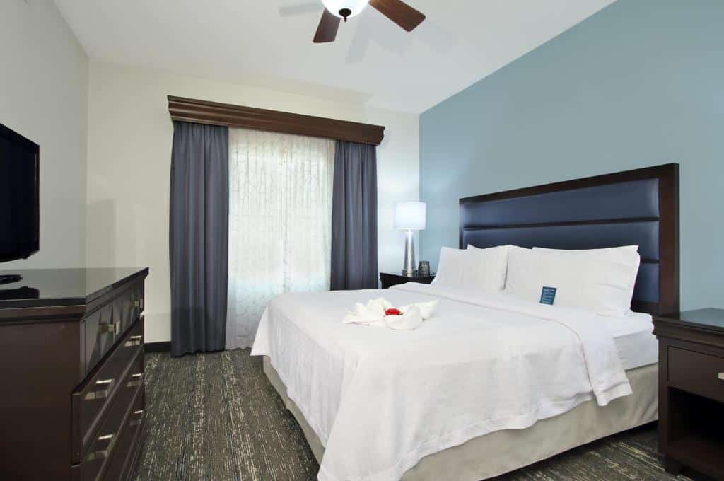 Quarto do Homewood Suites by Hilton Miami – Airport West com cama de casal, duas cômodas de madeira do lado da cama, uma cômoda de madeira com TV em frente da cama, do lado esquerdo janelas com cortinas brancas e roxas.