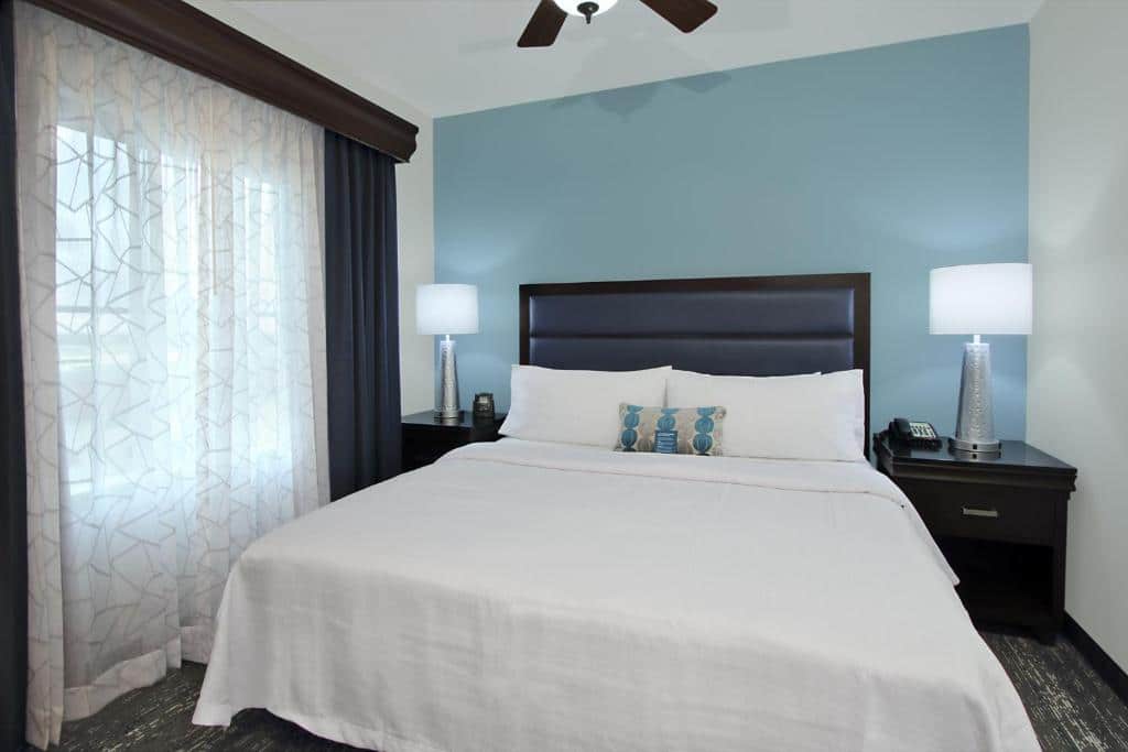 Quarto do Homewood Suites by Hilton Miami – Airport West com cama de casal, duas cômodas de madeira com luminária e janela com cortinas do lado esquerdo.