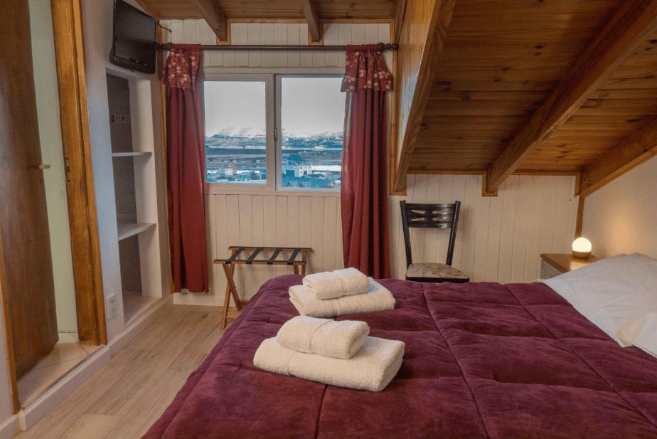 quarto do Hostería Aonikenk com uma cama de casal, colchas rosadas, toalhas brancas e uma pequena janela na parede com vista para as montanhas