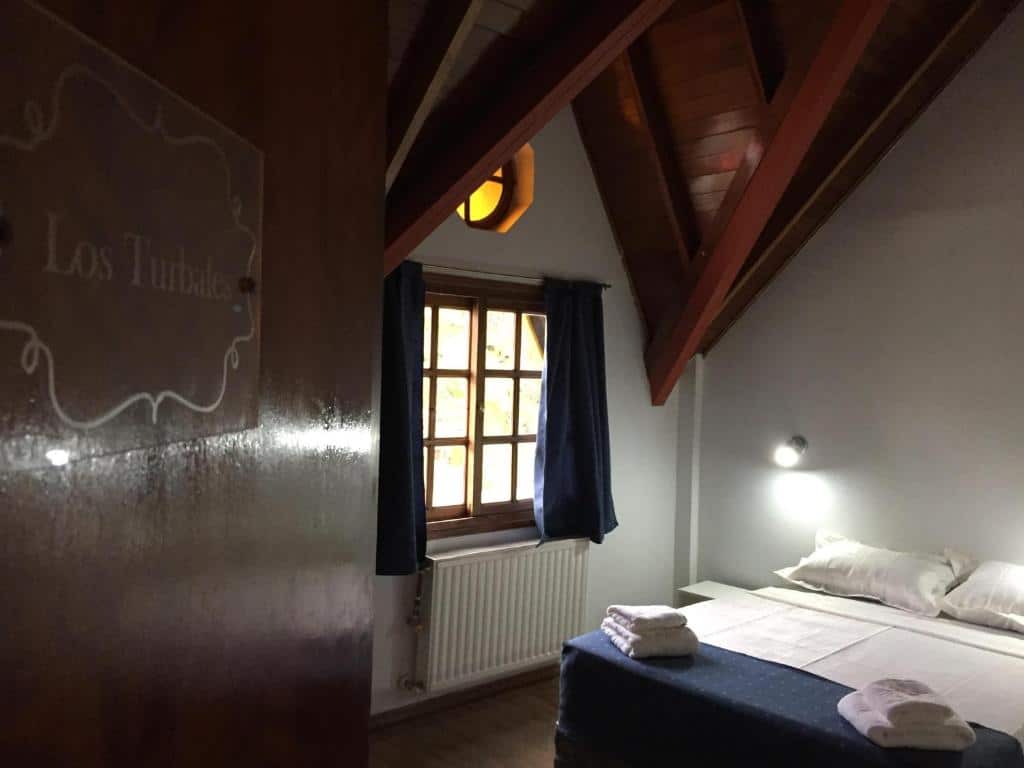 interior do quarto da Hosteria Kupanaka com uma cama de casal, uma janela pequena no lado esquerdo e teto catedral em madeira