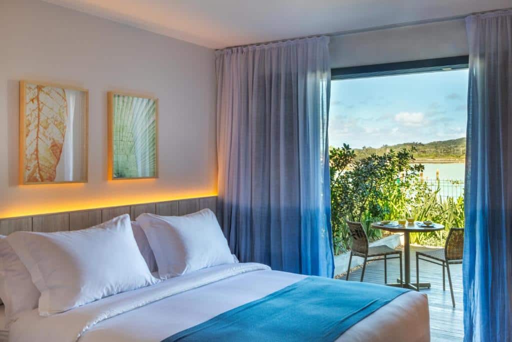 Quarto do Hotel Aretê  com uma sacada com cortinas e oferecendo vista para o mar, do lado de dentro é possível ver uma cama de casal com uma cabeceira com iluminação indireta