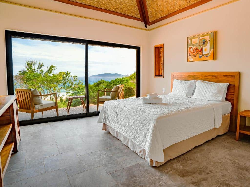 Quarto do Buzios Espiritualidade Hotel com uma cama aberta dando vista para o mar, com duas poltronas de madeira e, do lado de dentro, há um móvel de madeira e uma cama de casal