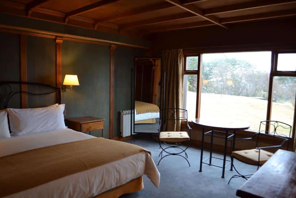 suíte do Hotel Los Ñires com uma cama de casal no canto esquerdo e uma peque área de descanso com uma mesa e duas cadeiras em frente a janela ampla com vista para a natureza